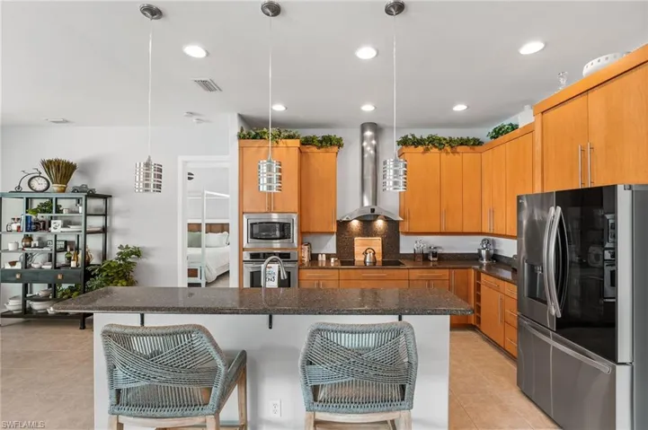 Kitchen featuring wall chimney range hood, a center island with sink, light tile patterned floors, stainless steel appliances, and decorative light fixtures