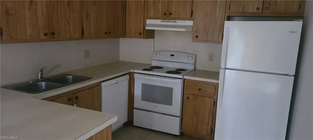 Kitchen featuring white appliances, light countertops, under cabinet range hood, light tile patterned floors, and brown cabinetry
