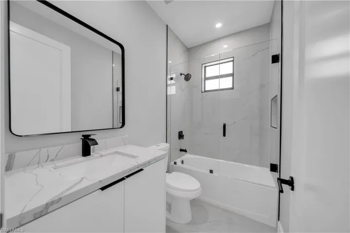 Modern bathroom featuring a single vanity with a white countertop and integrated sink, a rectangular framed mirror, a tub-shower combination with glass enclosure, white tile surround, and matte black fixtures
