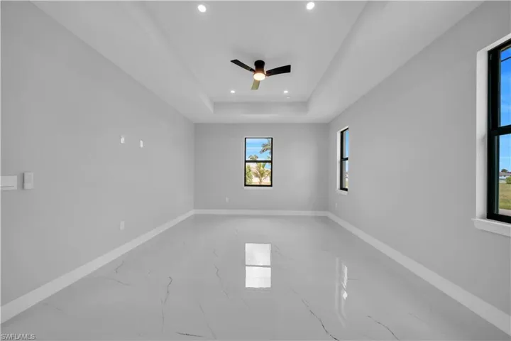 Spacious room featuring tray ceilings with recessed lighting, a ceiling fan, multiple windows with black framing, and large-format porcelain tile flooring