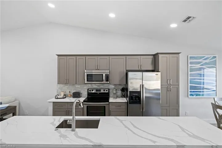 Kitchen featuring gray cabinets, stainless steel appliances, light stone countertops, vaulted ceiling, and decorative backsplash