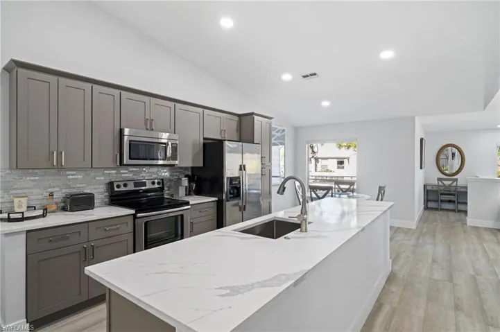 Kitchen featuring gray cabinetry, stainless steel appliances, vaulted ceiling, light wood-type flooring, and light stone counters