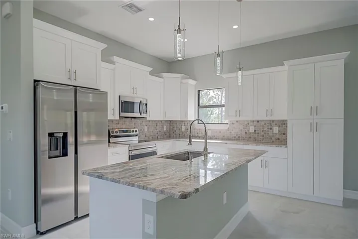 Kitchen with appliances with stainless steel finishes, a center island with sink, pendant lighting, light stone countertops, and white cabinetry