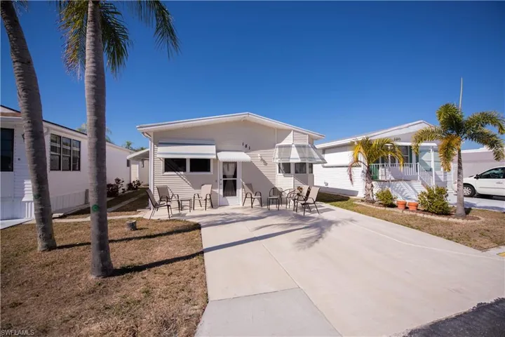 Single-story residence featuring light-toned horizontal siding, a concrete driveway, metal window awnings, and palm trees
