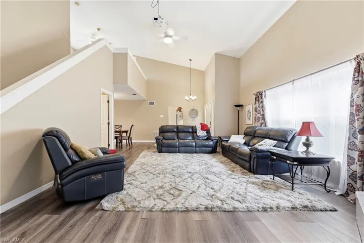 Vaulted ceiling living area with wood-finish flooring, a large window with sheers and curtains, and a staircase with a white railing