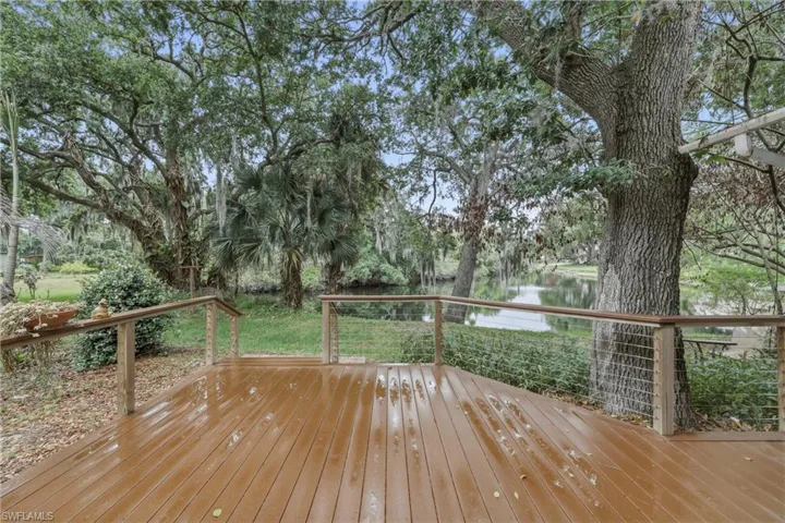 Expansive wood-finish deck with cable railing overlooking a tranquil waterway, bordered by mature trees and lush greenery