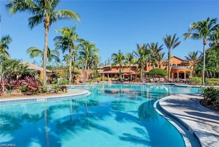 Expansive resort-style swimming pool featuring a curved design, surrounding tropical landscaping, and a terracotta-colored clubhouse with outdoor seating