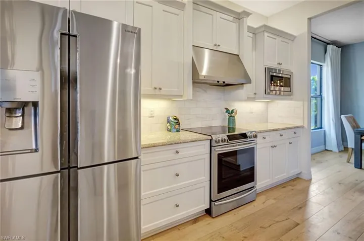 Kitchen with under cabinet range hood, light wood-style floors, appliances with stainless steel finishes, decorative backsplash, and light stone countertops