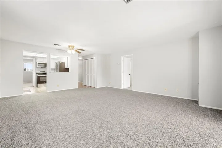 Spacious living area featuring light gray carpeting, neutral wall finishes, a ceiling fan with light fixture, and a pass-through window to the kitchen