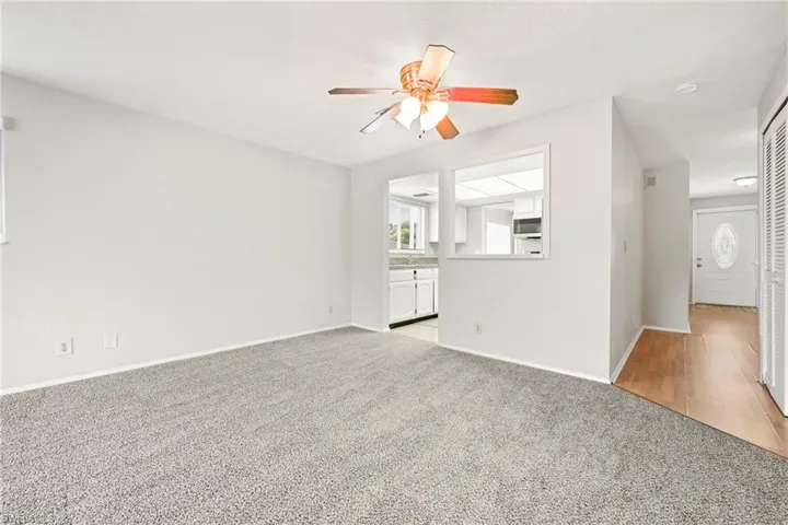 Carpeted living area featuring a ceiling fan with integrated lighting, a pass-through window to the kitchen, and a white paneled front entry door