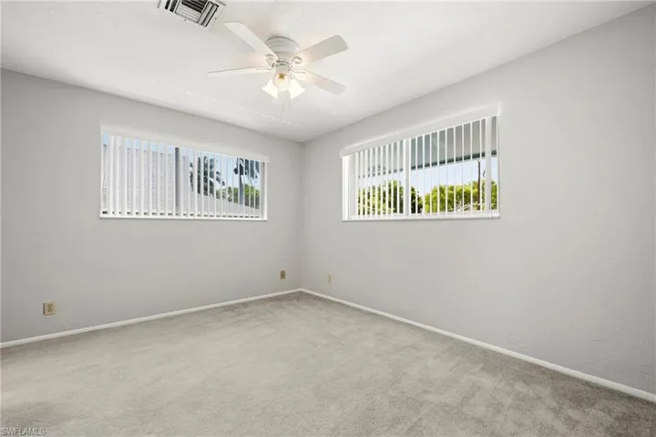 Spacious room featuring light gray carpet flooring, dual windows with vertical blinds, a ceiling fan with integrated lighting, and neutral wall paint