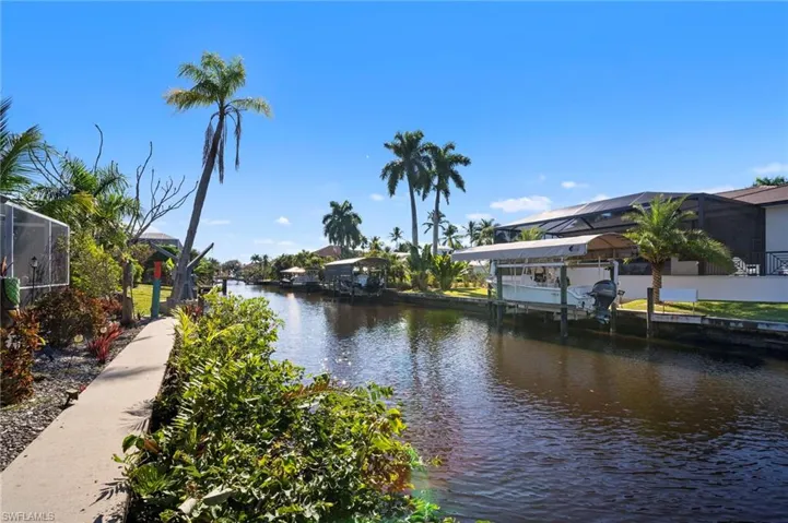 Waterfront property featuring mature palm trees, a concrete pathway, lush landscaping, and a boat lift with canopy