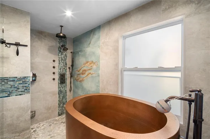 Bathroom featuring a copper soaking tub with floor-mounted bronzed fixtures, a tiled shower with multiple showerheads and body sprays, a mosaic tile accent wall, and a frosted window