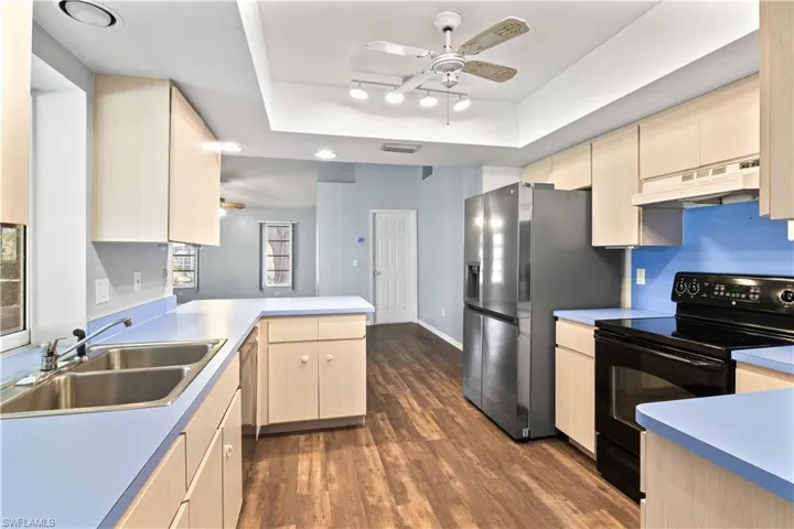 Kitchen with a raised ceiling, under cabinet range hood, black range with electric cooktop, light brown cabinets, and a sink