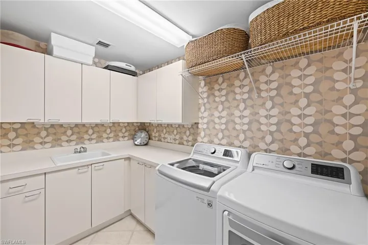 Dedicated laundry room featuring white cabinetry, geometric tile backsplash, top-load washer and front-load dryer with stainless steel finishes, woven storage baskets on floating shelving, and neutral tile flooring