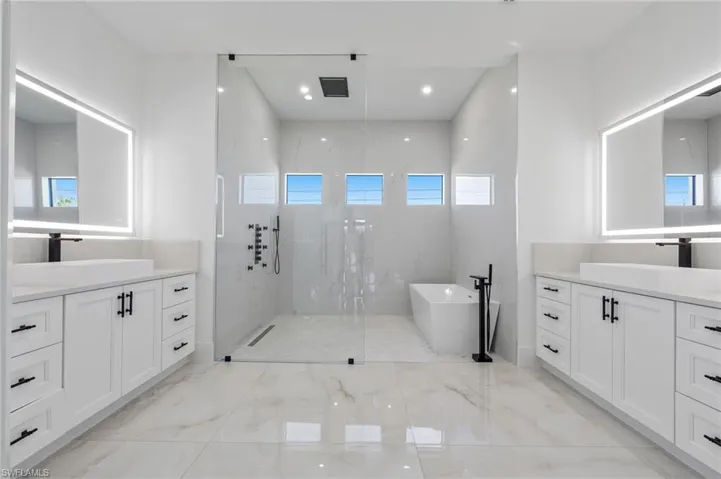 Dual white vanities featuring quartz countertops and illuminated mirrors, oversized walk-in shower with floor-to-ceiling tile, freestanding soaking tub, and matte black fixtures