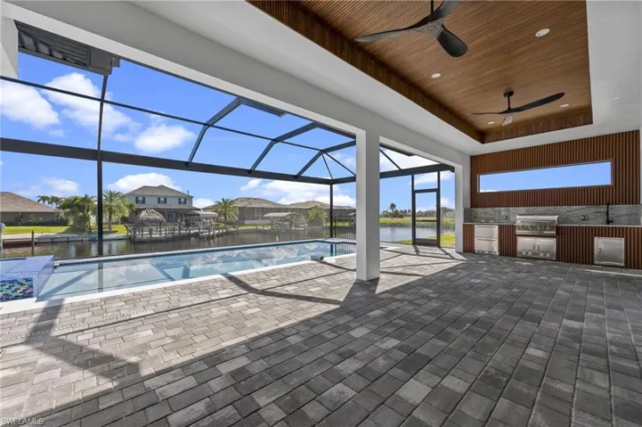 Covered outdoor living space featuring a wood-plank ceiling with dual ceiling fans, stone-paved flooring, built-in outdoor kitchen with stainless steel appliances, and a screened enclosure housing an in-ground pool