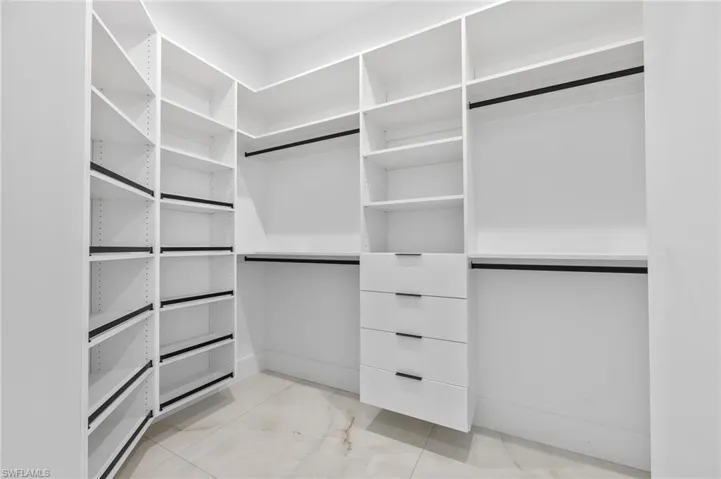 Custom walk-in closet featuring floor-to-ceiling white shelving, multiple hanging rods, four built-in drawers with matte black hardware, and stone-finish flooring