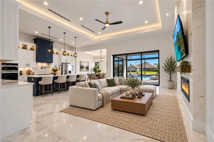 Open-concept living space featuring a tray ceiling with recessed lighting, tile flooring, built-in electric fireplace with stone-finish surround, kitchen island with contrasting cabinetry, and floor-to-ceiling glass sliding doors