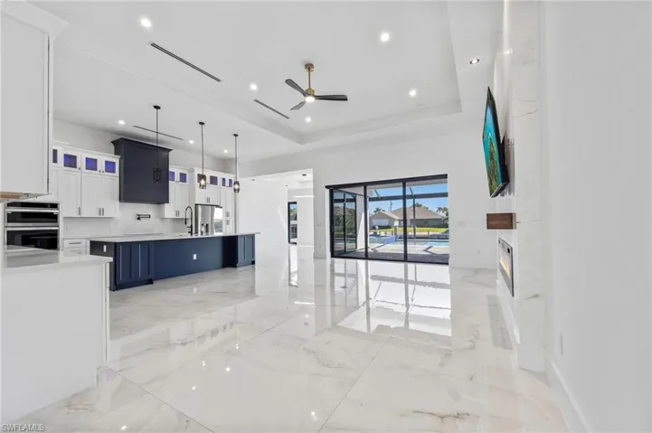 Open-concept interior featuring high tray ceilings, polished tile flooring, a kitchen island with dark-finish cabinetry, a built-in linear fireplace with stone-finish surround, and expansive sliding glass doors
