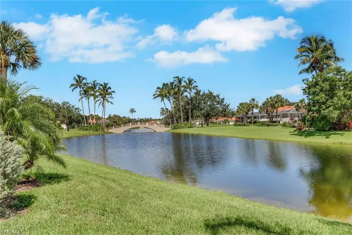 Expansive water feature with a pedestrian bridge, surrounded by mature palm trees, lush green landscaping, and distant tile-roof residences