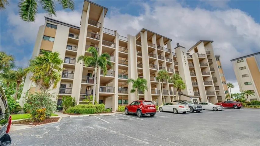 Multi-story building featuring multiple private balconies, a paved parking area, and mature palm trees