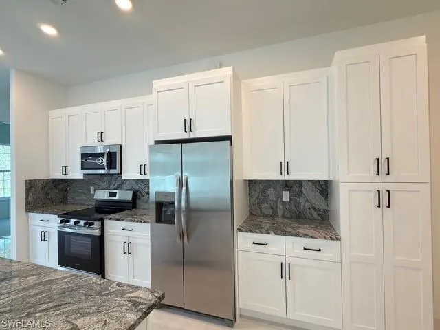 Kitchen with hanging light fixtures, stainless steel appliances, dark stone counters, white cabinetry, and decorative backsplash