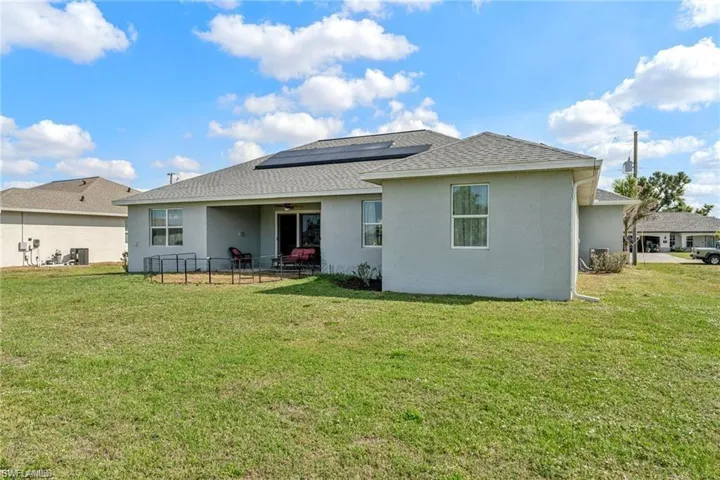 Rear view of property featuring stucco siding, solar panels, a ceiling fan, a shingled roof, and a lawn