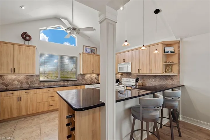 Kitchen with light brown cabinetry, backsplash, open shelves, decorative light fixtures, and high vaulted ceiling