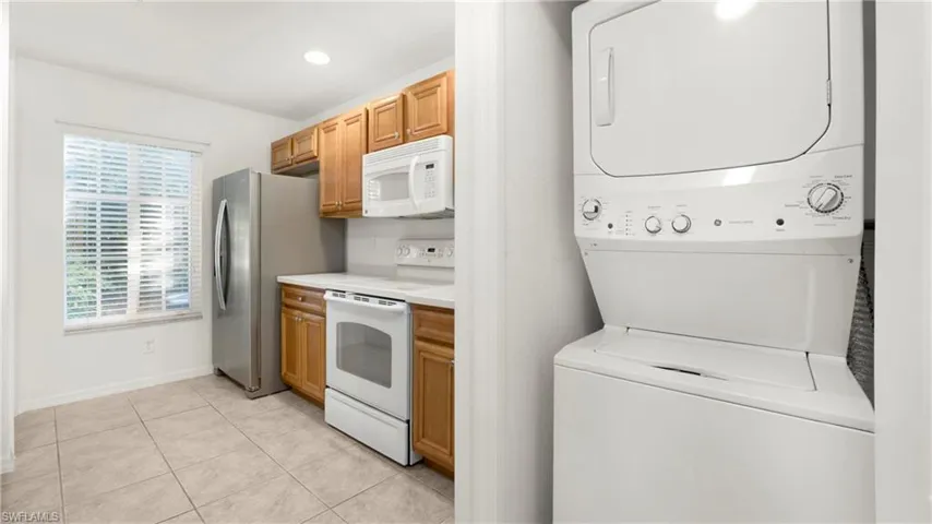 Kitchen with white appliances, light countertops, stacked washer and clothes dryer, light tile patterned floors, and brown cabinets
