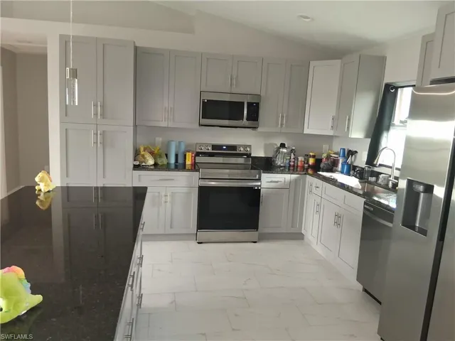 Kitchen with stainless steel appliances, dark stone countertops, light marble finish flooring, vaulted ceiling, and decorative light fixtures