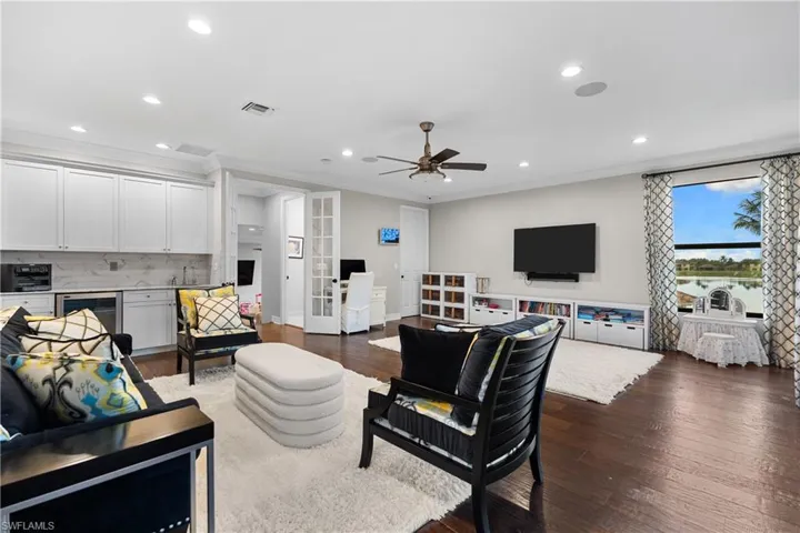 Spacious living area featuring rich wood-finish flooring, a contemporary wet bar with white cabinetry and a light-toned backsplash, recessed lighting, and an expansive window with outdoor water scenery