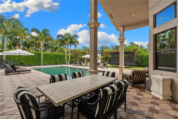 Expansive paver patio with a rectangular swimming pool, bordered by mature palm trees and a lush privacy hedge