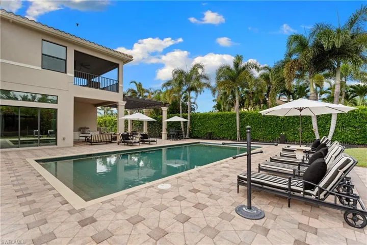 Expansive paver patio featuring a rectangular swimming pool, a second-story covered balcony with black railings, and a ground-level covered patio