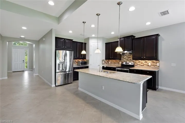 Kitchen featuring dark wood cabinetry, stainless steel appliances, a multi-tone tile backsplash, and an island with a light-toned countertop