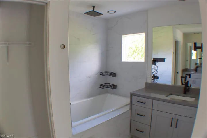 Bathroom featuring a soaking tub, wall-mounted showerhead, marble-look wall tiling, and a light gray vanity with a single basin sink and dark hardware
