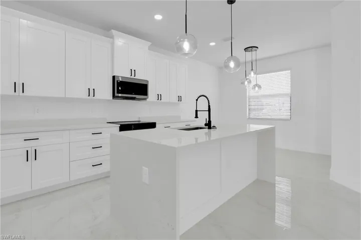 Kitchen featuring light stone countertops, white cabinetry, an island with sink, stainless steel microwave, and decorative light fixtures