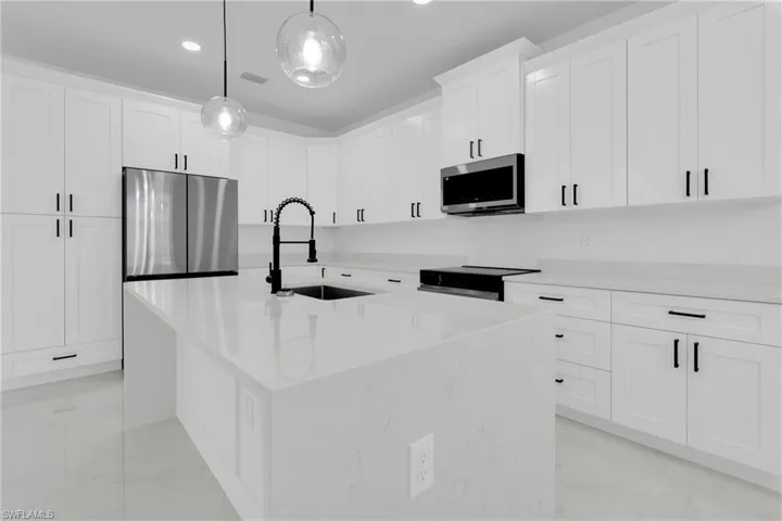 Kitchen featuring stainless steel appliances, light stone countertops, an island with sink, white cabinetry, and hanging light fixtures