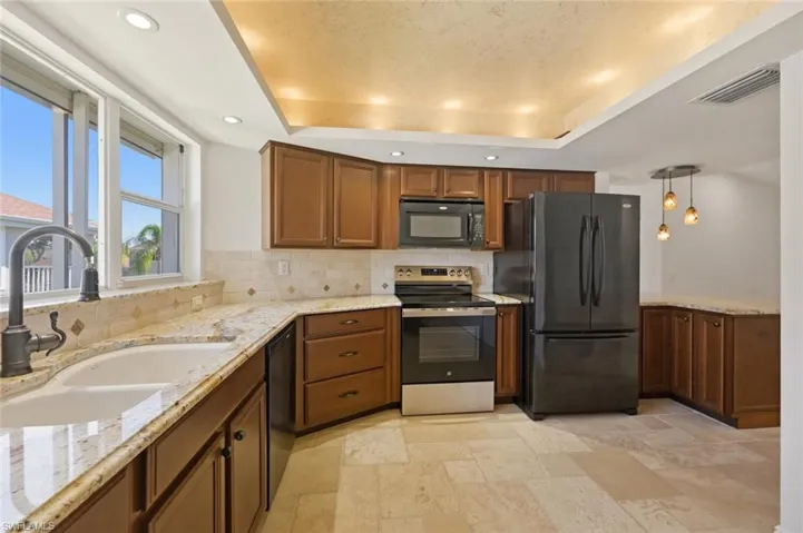 Kitchen with a raised ceiling, black appliances, light stone counters, pendant lighting, and wood finish cabinetry