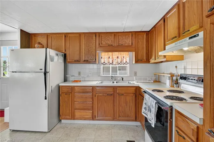 Kitchen featuring white appliances, under cabinet range hood, light countertops, brown cabinetry, and tasteful backsplash