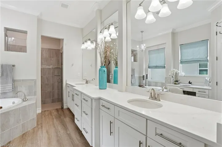 Dual vanity with white cabinetry and integrated sinks set beneath large mirrors, wood-finish flooring, a tiled corner soaking tub, and a shower with glass enclosure
