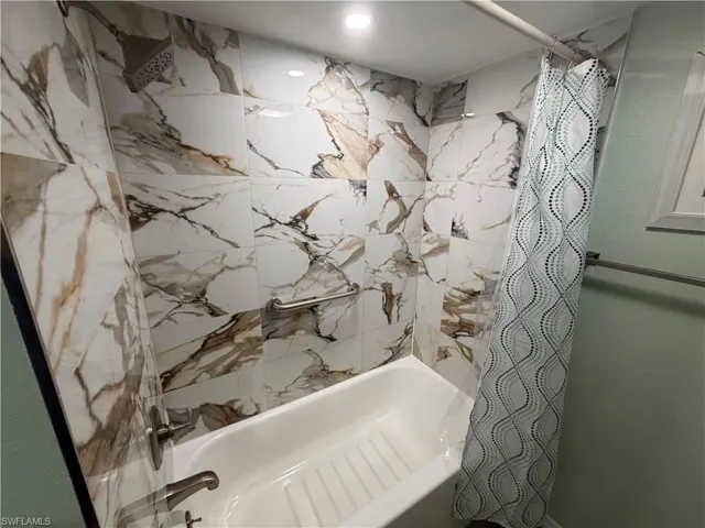 Bathtub with white and brown veined tile surround, brushed nickel showerhead, and wall-mounted grab bar