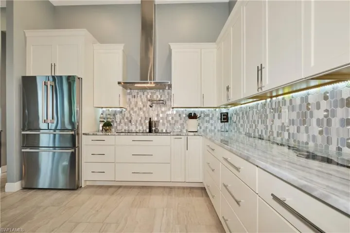 Kitchen with wall chimney exhaust hood, tasteful backsplash, white cabinetry, light stone countertops, and stainless steel fridge