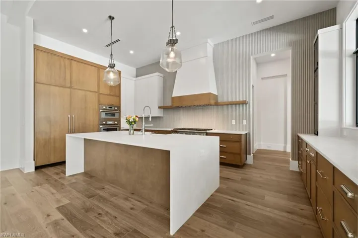 Kitchen with light wood-type flooring, pendant lighting, a kitchen island with sink, tasteful backsplash, and custom range hood