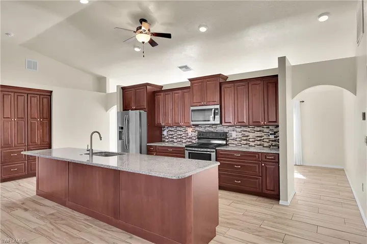 Kitchen with arched walkways, appliances with stainless steel finishes, light stone counters, a kitchen island with sink, and vaulted ceiling