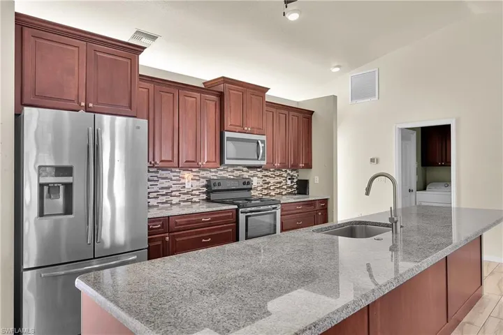 Kitchen featuring appliances with stainless steel finishes, light stone counters, washer / dryer, vaulted ceiling, and an island with sink