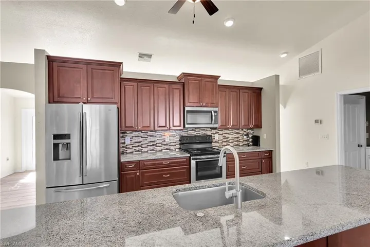 Kitchen featuring appliances with stainless steel finishes, light stone countertops, arched walkways, reddish brown cabinets, and decorative backsplash