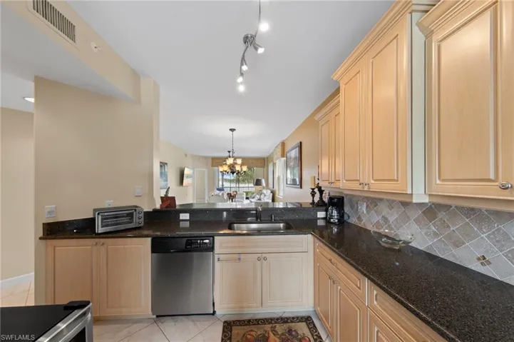Kitchen featuring stainless steel dishwasher, suspended lighting, dark stone counters, light wood finish cabinets, and backsplash