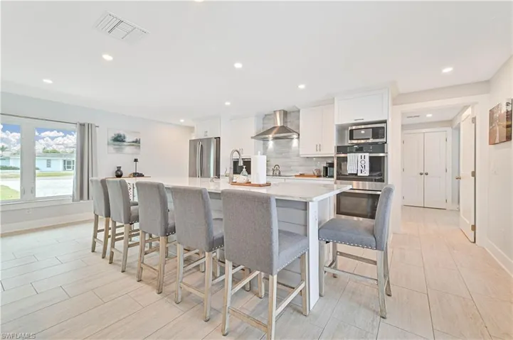 Kitchen with stainless steel appliances, wall chimney exhaust hood, a kitchen island with sink, white cabinets, and a breakfast bar