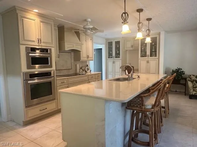 Kitchen with decorative light fixtures, premium range hood, tasteful backsplash, a kitchen island with sink, and stainless steel double oven